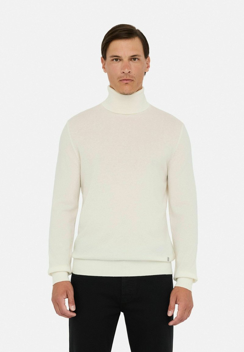 Mann in einem enganliegenden weißen Rollkragenpullover und schwarzen Hosen, der vor einem schlichten weißen Hintergrund steht.