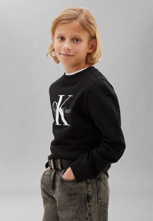 Lyshåret barn iført en sort Calvin Klein sweatshirt og grå jeans med hænderne i lommerne, stående mod en ensartet lysegrå baggrund.