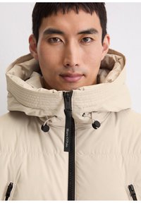 Beige donsjack met een hoge kraag, ritssluiting en trekkoorddetails. Gestructureerde stof met een glanzende afwerking en zijzakken.