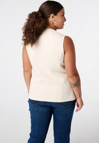 Lichtbeige mouwloos topje met een getailleerd ontwerp, textuurstof en zichtbare naden, gecombineerd met donkerblauwe denim broek.