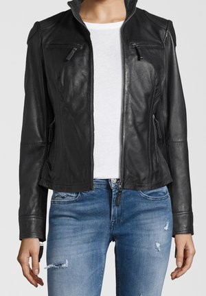 Femme portant une veste en cuir noire sur une chemise blanche et un jean bleu déchiré, debout les bras détendus le long du corps.