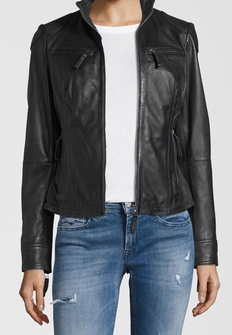 Femme portant une veste en cuir noire sur une chemise blanche et un jean bleu déchiré, debout les bras détendus le long du corps.