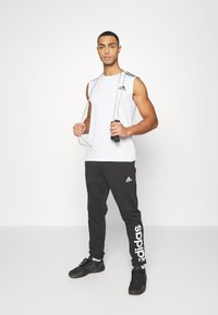Sportisks apģērbs, kas sastāv no baltas bezpiedurkņu krekla ar melniem akcentiem un logo, apvienojot to ar melniem sporta biksēm un melniem sporta apaviem.