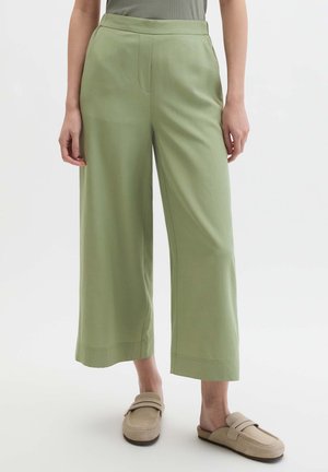 Osoba ubrana w jasnozielone szerokie spodnie o długości do połowy łydki oraz beżowe wsuwane mokasyny, stojąca na tle jednolitego białego tła.