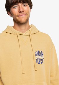 Yellow cotton hoodie with a front pocket, drawstring hood, and circular blue graphic logo featuring text on the chest. Soft texture.