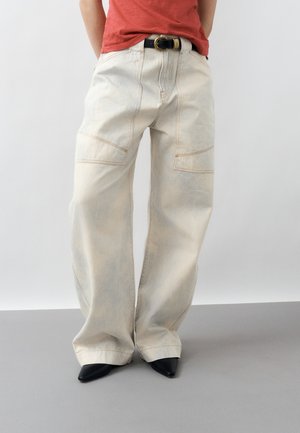 Άτομο με εκρού παντελόνι ευρύ σε φαρδιά γραμμή με ορατές λεπτομέρειες ραφής, μαύρα μυτερά παπούτσια, μαύρη ζώνη και κόκκινη μπλούζα μέσα στην παντελόνα.