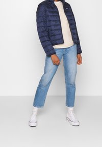 Personne portant une doudoune navy, un pull crème, un jean bleu clair, des chaussettes blanches et des baskets montantes blanches, se tenant contre un fond blanc.