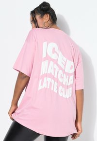 Kobieta z kolczykami w kształcie kół nosi dużą różową koszulkę z napisem "ICED MATCHA LATTE CLUB" w dużych białych literach na plecach.