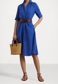 Blå linnaklänning med korta ärmar, åtdragen i midjan med ett brunt bälte. Kompletterad med en vävd stråväska och bruna sandaler.