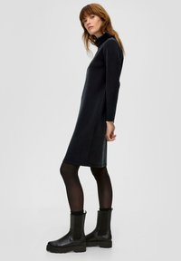 Czarna sukienka z długim rękawem, z wysokim kołnierzem, prostym krojem i bocznymi rozporkami, zestawiona z czarnymi botkami za kostkę z elastycznymi panelami bocznymi.