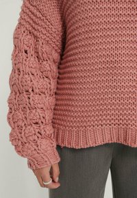 Stickad tröja i dammros, med texturerad, tjock garn och utsmyckade, öppna ärmar. Ribbat fåll och manschetter ger visuell intresse.