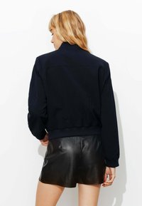 Veste de bombardier marine avec des poignets et un ourlet côtelés, présentant une texture lisse et une coupe décontractée. Assortie à des shorts en cuir noirs.