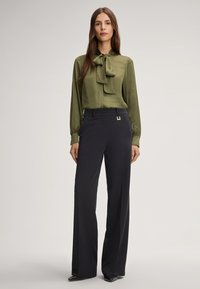 Blusa di raso verde con fiocco al collo, abbinata a pantaloni a gamba larga neri a vita alta con un accento metallico.