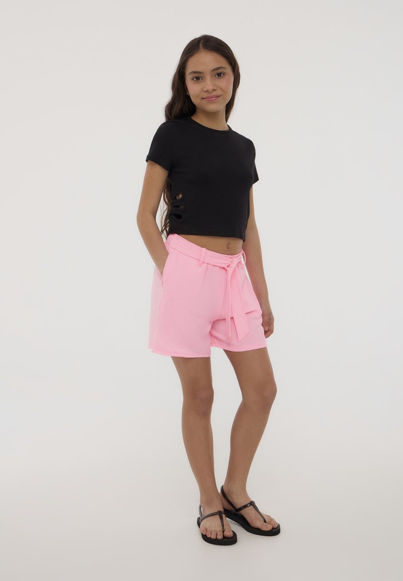 Maglietta nera corta con tagli laterali, abbinata a pantaloni corti rosa con texture e cordino in vita. Il modello indossa infradito nere, in piedi su uno sfondo chiaro.