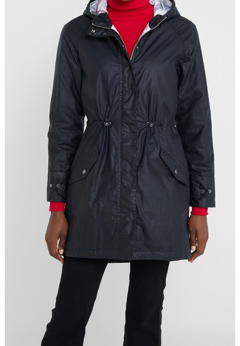 Femme portant un imperméable noir à capuche avec poches avant et taille cintrée, par-dessus un col roulé rouge et un pantalon noir, debout devant un fond uni.