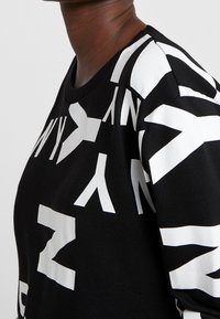 Persoon draagt een zwart shirt met lange mouwen, bedrukt met grote, gedurfde witte letters en geometrische vormen over de borst en mouwen.