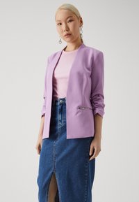 Lilac-takk jakke med skreddersydd passform, åpen front og rynkede ermer; kombinert med en ribbestrikket rosa topp og en blå denimskjørt med splitt foran.