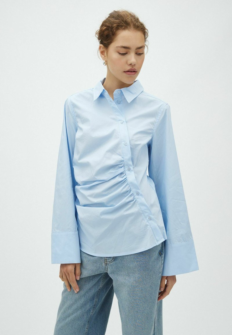 Femme portant une chemise bleu clair à manches longues avec des détails froncés et de larges poignets, associée à un jean bleu clair, regardant vers le bas.