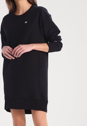 Femme portant une robe-sweat à manches longues noire de la marque Champion avec des poignets côtelés et un petit logo sur la poitrine, debout devant un fond uni.