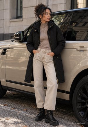 Une femme portant un manteau noir matelassé, un haut rayé, un jean clair et des bottes s’appuie contre un SUV beige dans une rue de la ville.