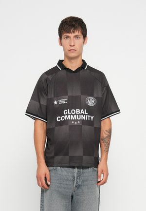 Bărbat îmbrăcat cu o cămașă scurtă cu mâneci în carouri negre și gri, cu textul "Global Community", asortată cu blugi din denim albastru deschis, stând în fața unui fundal alb.