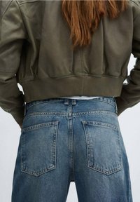 Chaqueta bomber verde oliva con dobladillo acanalado, combinada con jeans de denim azul de cintura alta que cuentan con dos bolsillos traseros y una textura ligeramente desgastada.