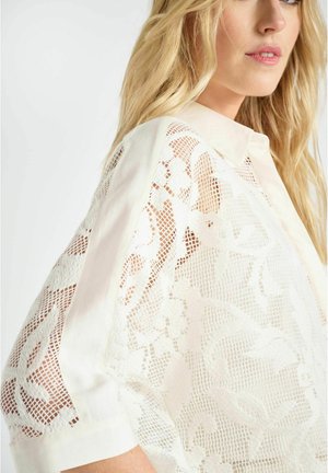 Femme blonde portant un chemisier en dentelle blanche avec des motifs floraux et des panneaux en tulle transparent, vue en gros plan de profil latéral.