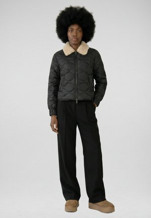 Personne noire avec une grande afro portant une veste matelassée noire avec un col beige, un pantalon large noir et des chaussures à plateforme beige, debout sur fond gris.