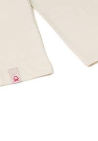 Krémový žebrovaný top s dlouhým rukávem a čtvercovým střihem; na dolním okraji je růžová etiketa s grafickým detailem. Měkká, texturovaná tkanina.