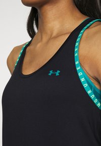Zbliżenie na osobę w czarnym sportowym topie na ramiączkach z turkusowym logotypem Under Armour i turkusowymi ramiączkami z napisem "Under Armour".