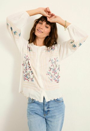 Kvinde iført hvid bluse med blomsterbroderi og puffede ærmer, kombineret med lyseblå jeans, poser med hænderne over hovedet.