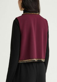 Blouse sans manches bordeaux avec une base à manches longues noire, présentant une bordure texturée dorée et métallique le long de l'ourlet. Finition en tissu lisse.