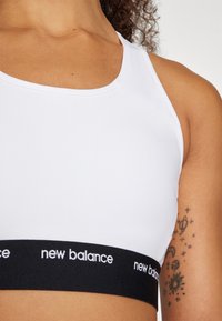 Balta sportinė liemenėlė su apvaliu iškirpimu ir juodu elastingu juostos su užrašu "new balance", atspausdintu balta spalva. Lygus paviršius ir prigludusi forma.