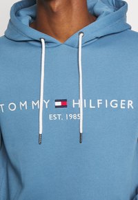Světle modrá mikina s kapucí a designem na stahování, bílý nápis "TOMMY HILFIGER EST. 1985" a logo s červenou, bílou a modrou barvou.