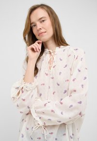Blusa bianca con fantasie paisley in rosso e blu. Presenta un colletto da legare e maniche a palloncino elasticizzate con dettagli arricciati. Tessuto leggero.