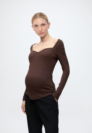 Femme enceinte aux cheveux blonds porte un haut marron côtelé à manches longues et un pantalon noir, debout avec une main dans la poche sur fond uni.