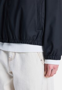 Ναυτικό μπουφάν αδιάβροχο με ελαστικές μανσέτες, σε συνδυασμό με λευκή ριπ υφής τρέσσα. Ορατές ραφές σε ανοιχτόχρωμο παντελόνι, χέρι με τατουάζ κοντά στο τελείωμα.