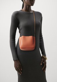 Piccola borsa a tracolla in pelle arancione con base arrotondata, texture liscia e tracolla regolabile, indossata con un vestito grigio a coste.