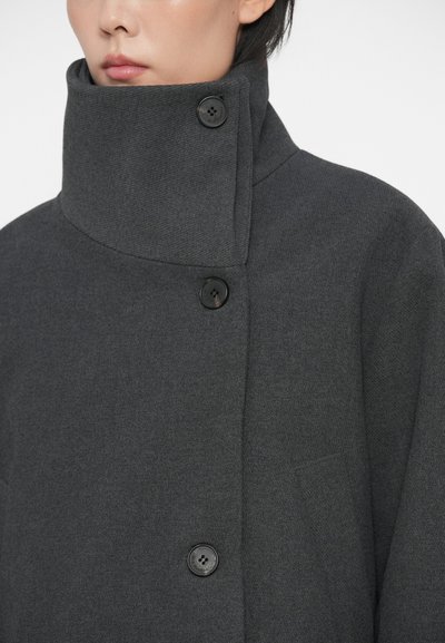 Person iført en mørkegrå højhalset frakke fastgjort med tre runde sorte knapper, delvist synlige læber og hage.