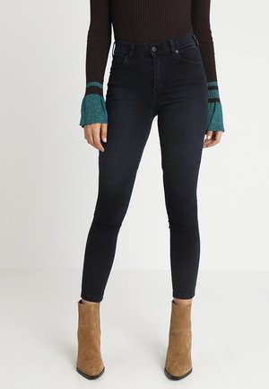 Femme portant un jean skinny noir taille haute, un pull côtelé marron avec des poignets bleu sarcelle, et des bottines en suède beige sur fond blanc.