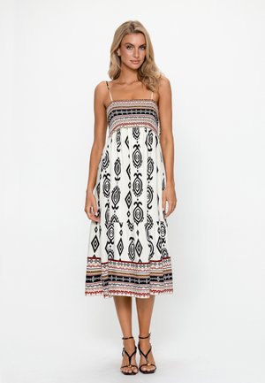 Frau trägt ein ärmelloses weißes Midikleid mit schwarzen geometrischen und roten gemusterten Designs, steht mit entspannten Händen an den Seiten.