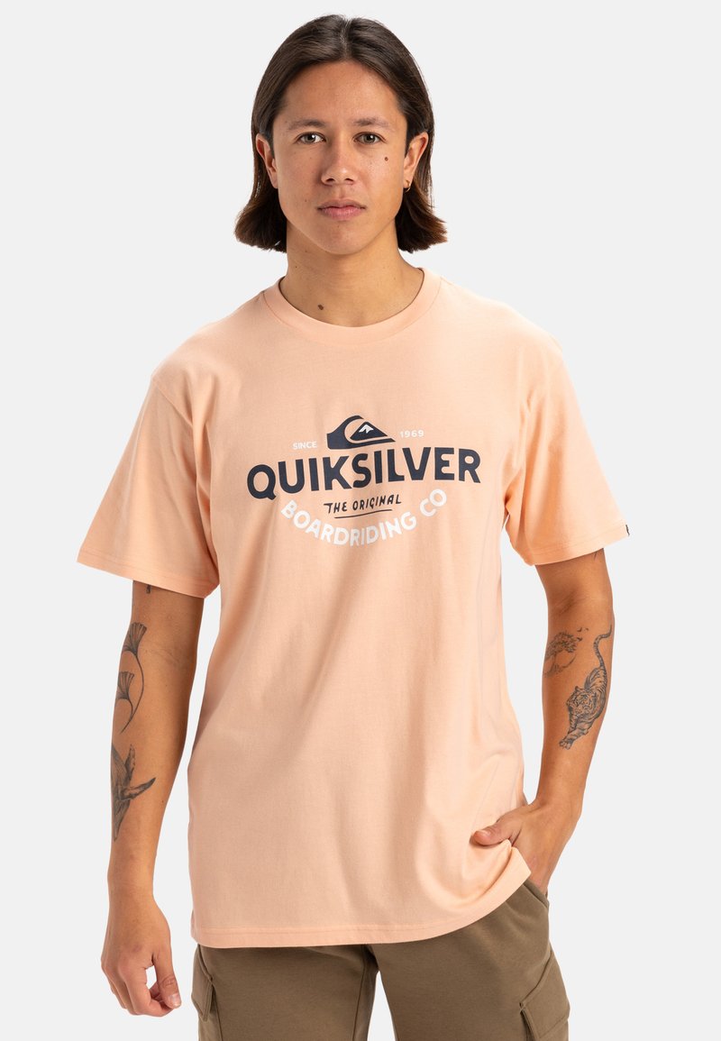 Camiseta de algodón color melocotón claro con un logo de Quiksilver en azul oscuro, corte relajado, cuello redondo y mangas cortas.