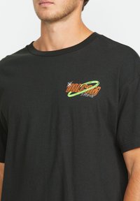Schwarzes Baumwoll-T-Shirt mit einem Front-Graphic, das den orangen und grünen Text "Vulkanstein" in einem runden Design-Stil zeigt. Kurze Ärmel.