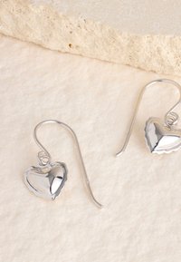 Silberne herzförmige Ohrringe mit polierter Oberfläche und wellenförmigen Kanten, ausgestattet mit einer Fischhakenbefestigung. Strukturierter Hintergrund.