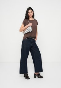 Καφέ μπλούζα με κοντά μανίκια συνδυασμένη με φαρδιά σκουρόχρωμα τζιν παντελόνια, μια μαύρη ζώνη με μεταλλικές λεπτομέρειες και λαμπερά μαύρα παπούτσια με τακούνι.