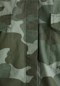 Tela de camuflaje verde y gris con costuras cosidas y solapas de bolsillo, que muestra un primer plano de la sección frontal de una prenda.