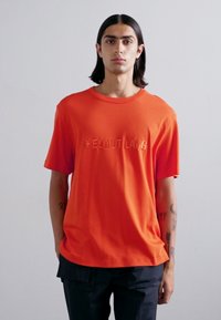 Camiseta de algodón naranja con logotipo de "HELMUT LANG" en relieve, cuello redondo, mangas cortas y un ajuste relajado, combinada con pantalones oscuros.