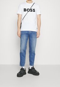 Valkoinen T-paita, jossa musta "BOSS" -painatus, siniset farkut ja mustat lenkkarit, joissa on heijastavat yksityiskohdat ja valkoiset sukat. Yksinkertainen muotoilu, rento istuvuus.