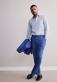 Muž v modrej kvetinovej košeli a modrých nohaviciach drží zodpovedajúce modré sako, stojí s prekríženými nohami pri jednofarebnej stene a kobercovej podlahe.