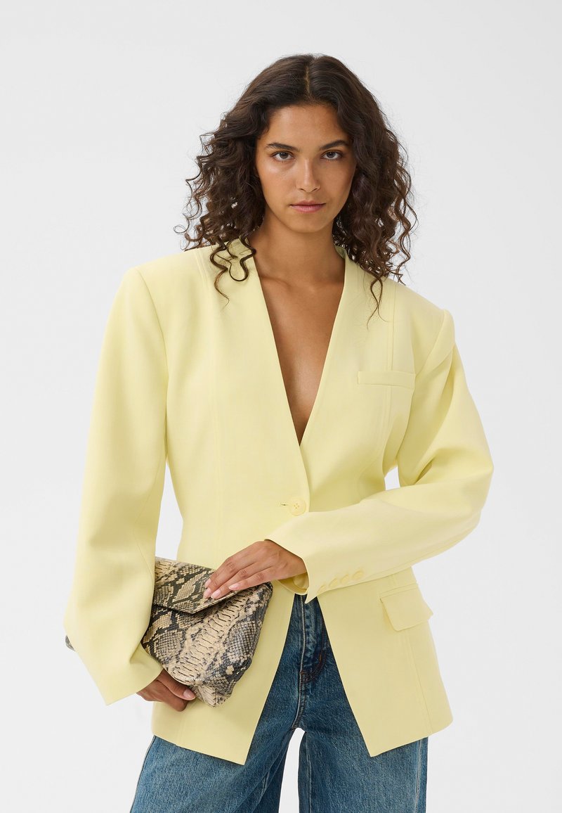 Femme aux cheveux bouclés portant un blazer oversize jaune pâle et un jean bleu, tenant une grande pochette à motif peau de serpent.